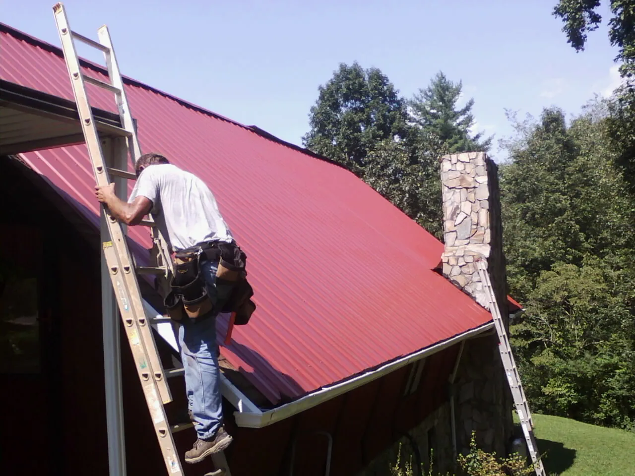 Roofer on a ladder beside a red metal roof and stone chimney — Soffit & Fascia Repair by Roofing Brothers in Bensalem
